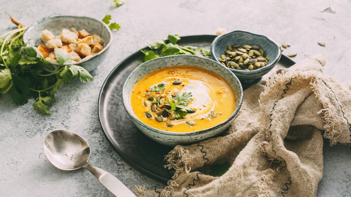 Eine Schüssel Kürbis-Curry-Suppe mit Kernen, Croûtons und Kräutern verfeinert, daneben je eine Schüssel mit Kernen und Croûtons.