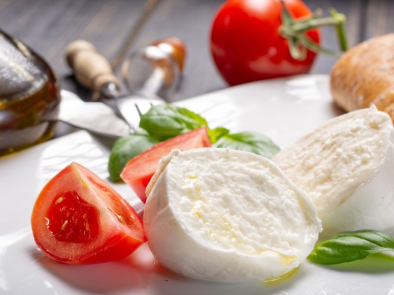 Eine halbierte, selbstgemachte Mozzarella-Kugel auf einem Teller. Daneben Tomaten, Basilikum und Brot.