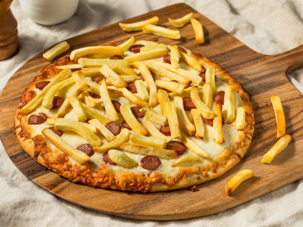 Eine Pizza Americana mit Pommes und Wiener Würstchen auf einem Holzbrett.