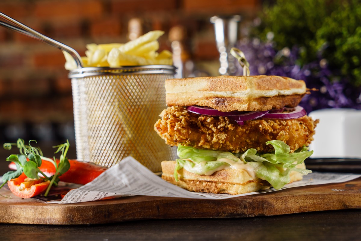 Ein Waffel-Burger belegt mit Hähnchen, Salat, Gewürzgurke und roter Zwiebel, daneben ein Korb mit Pommes.