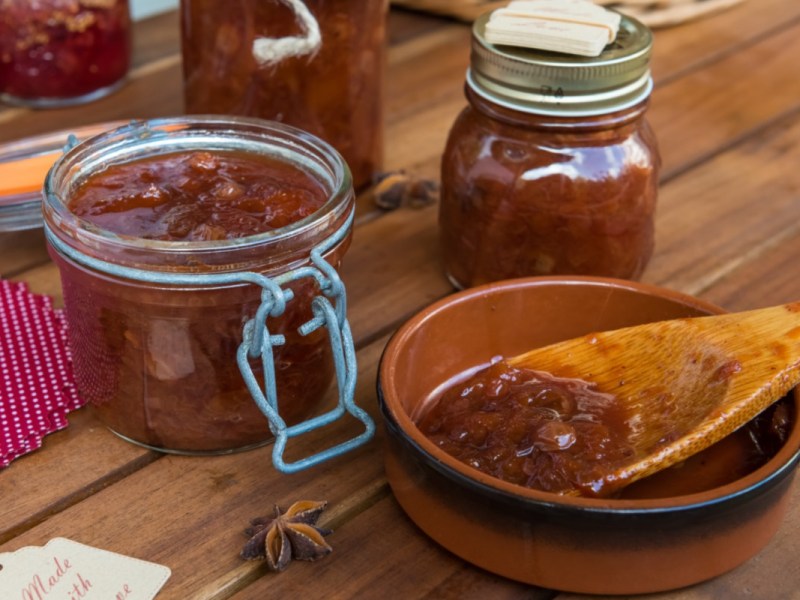 Mehrere Gläser Zwetschgen-Chitney. Daneben ein Holzlöffel mit Chutney, der in einer kleinen Schale liegt.