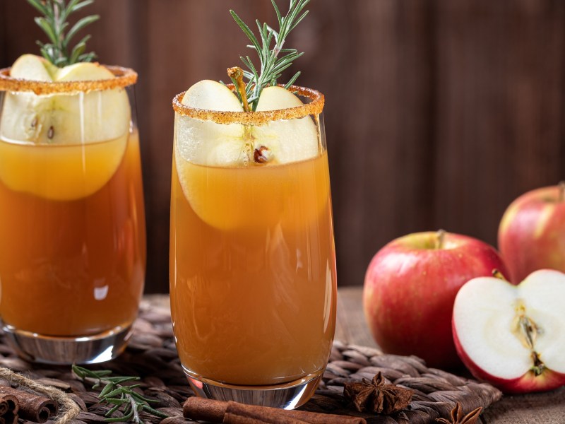 Herbst-Cocktail in zwei Gläsern, garniert mit Apfel und Rosmarin. Daneben halbierte und ganze Äpfel.