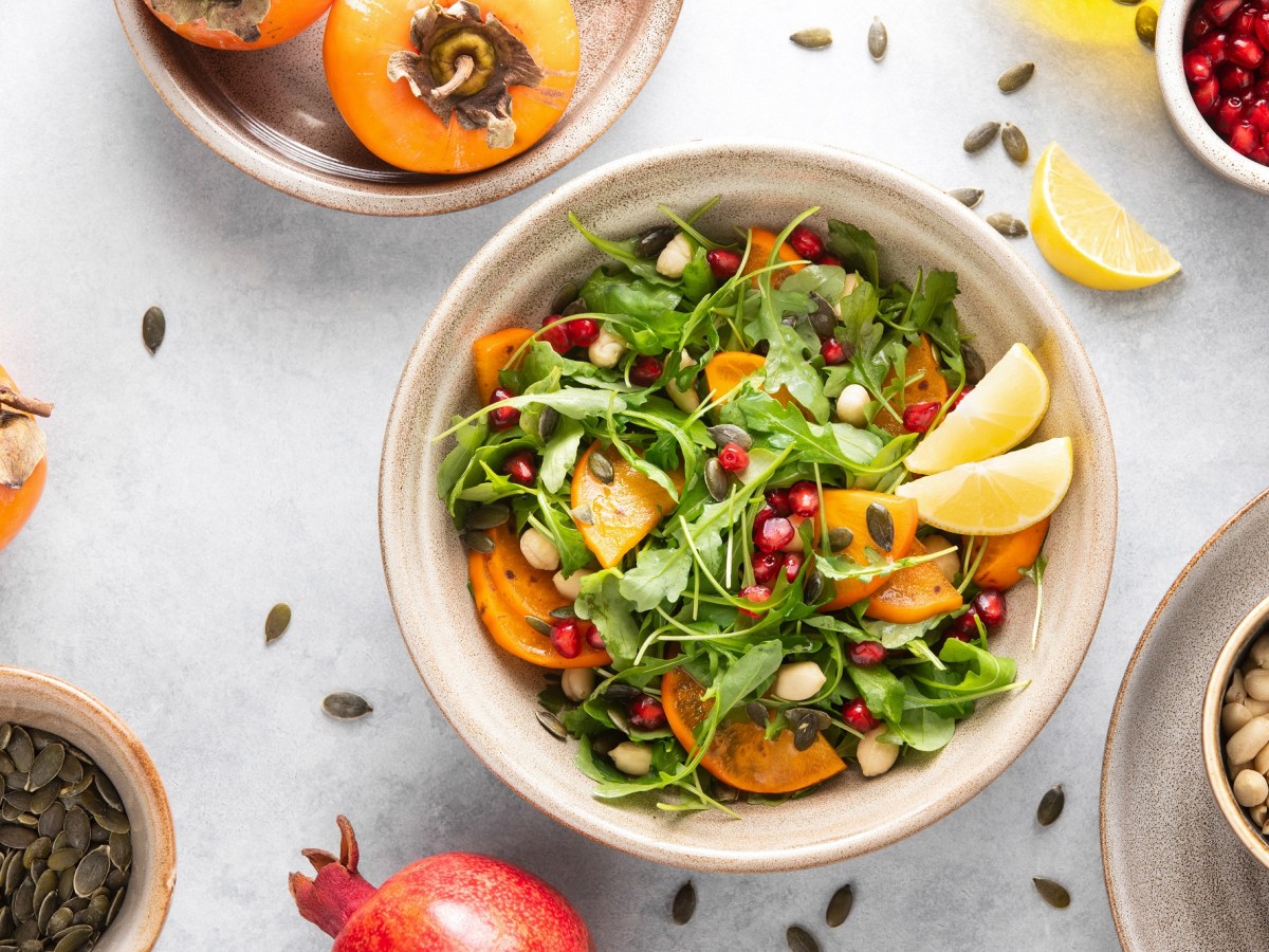 Kaki-Salat mit Rucola in einer weißen Schale. Daneben liegen ein Granatapfel, Nüsse und Kerne.