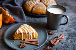 Ein Kürbis-Scones verziert mit Zuckerguss auf einem Teller, daneben Zimtstangen und eine Tasse Milch, im Hintergrund weitere Scones und Kürbisspalten.