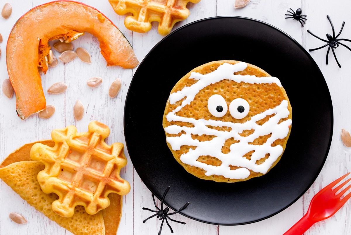 Mumienpfannkuchen mit Zuckerguss auf einem schwarzen Teller, umgeben von Halloween-Dekoration.