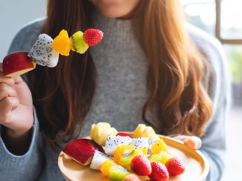 Obst am Abend essen: Eine Frau hält einen Obstspieß in der rechten und einen Teller mit Obstspießen in de rlinken Hand.