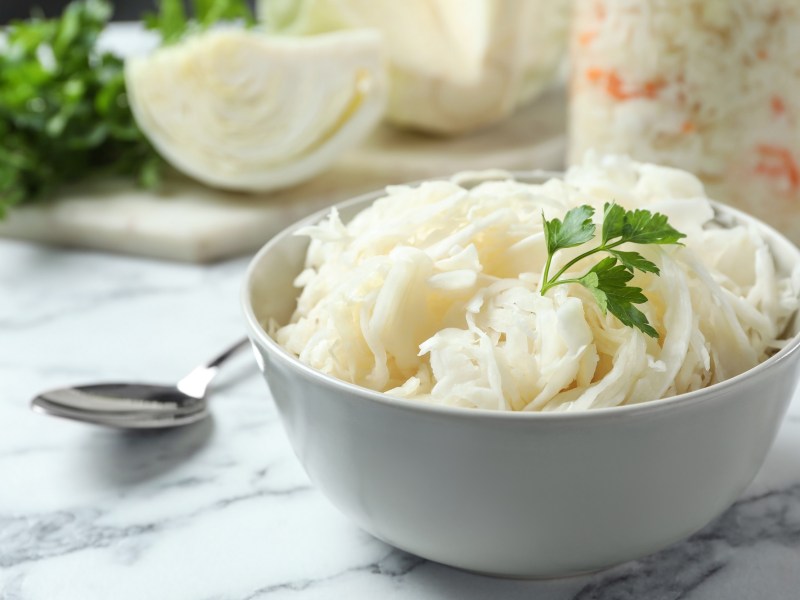 Eine Schüssel südtiroler Krautsalat, daneben ein Löffel, im Hintergrund ein Glas mit dem Salat und ein Kohlstück.