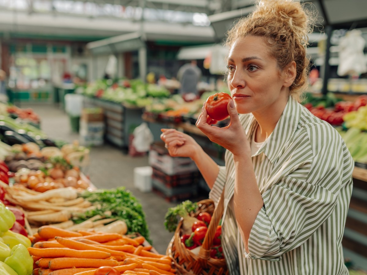 Vegane Mythen: eine Frau steht am Gemüsestand und hält eine Tomate in der einen und einen Einkaufskorb in der anderen Hand.