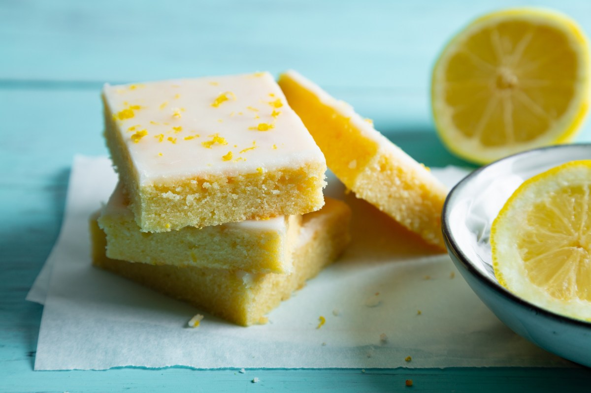 4 Stücke Zitronen-Brownies Lemonies auf Backpapier, daneben halbierte Zitronen.