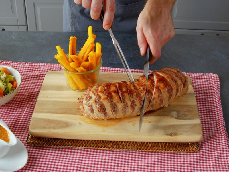 Ein gefüllter Schweinebraten wird mit einem Messer angeschnitten. Daneben Maispommes.