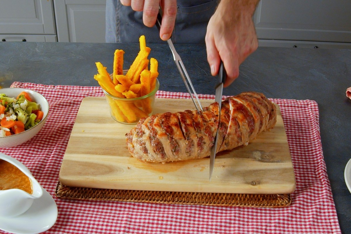 Ein gefüllter Schweinebraten wird mit einem Messer angeschnitten. Daneben Maispommes.