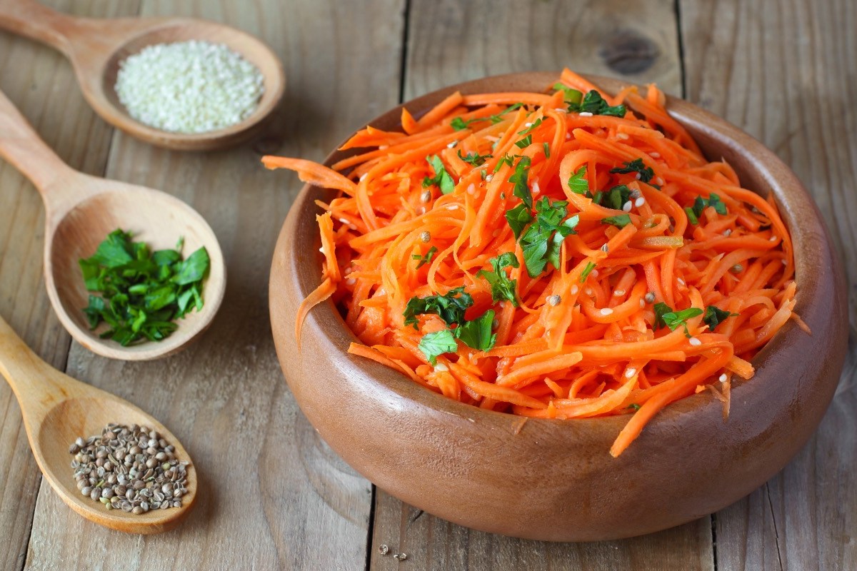 Eine Schüssel koreanischer Karottensalat. Daneben liegen Holzlöffel mit Gewürzen.