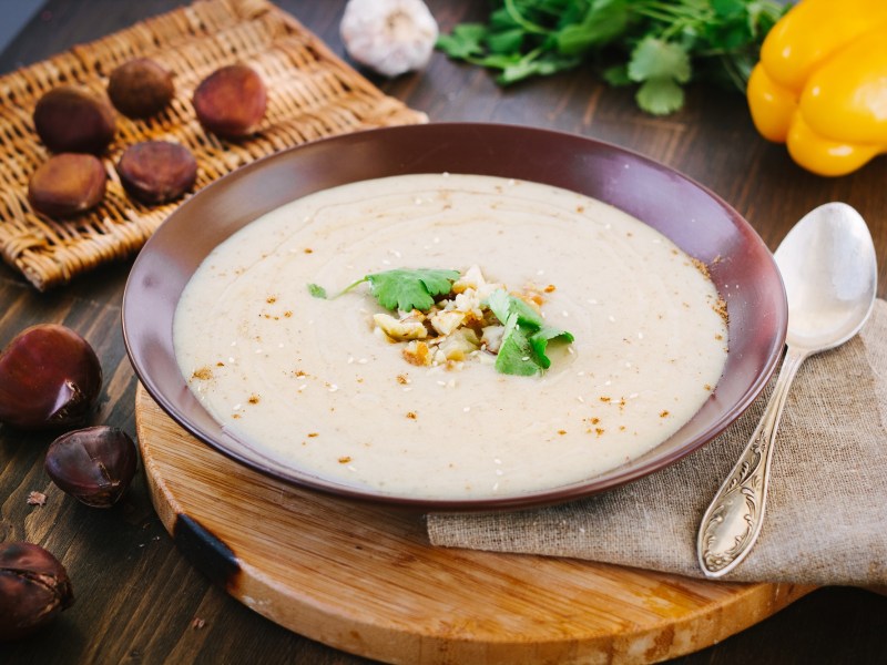 Ein Teller Maronensuppe mit BergkÃ¤se, daneben ein LÃ¶ffel und Maronen.