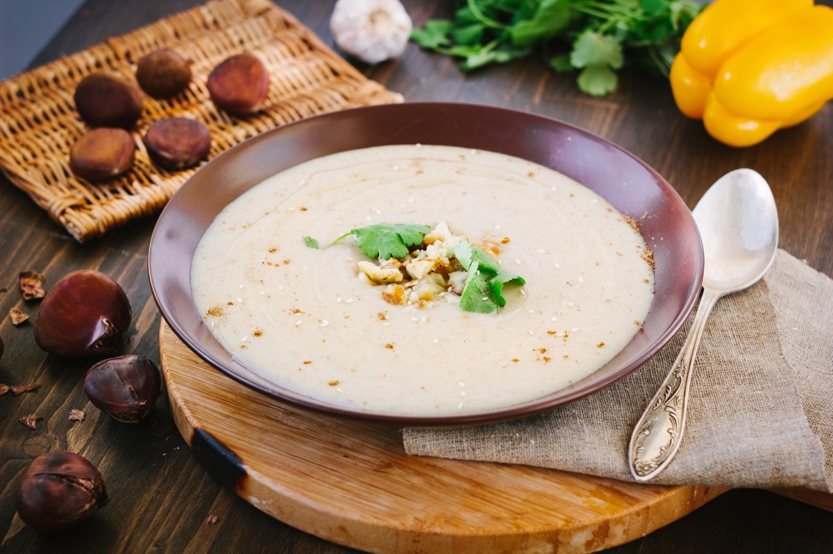 Ein Teller Maronensuppe mit BergkÃ¤se, daneben ein LÃ¶ffel und Maronen.