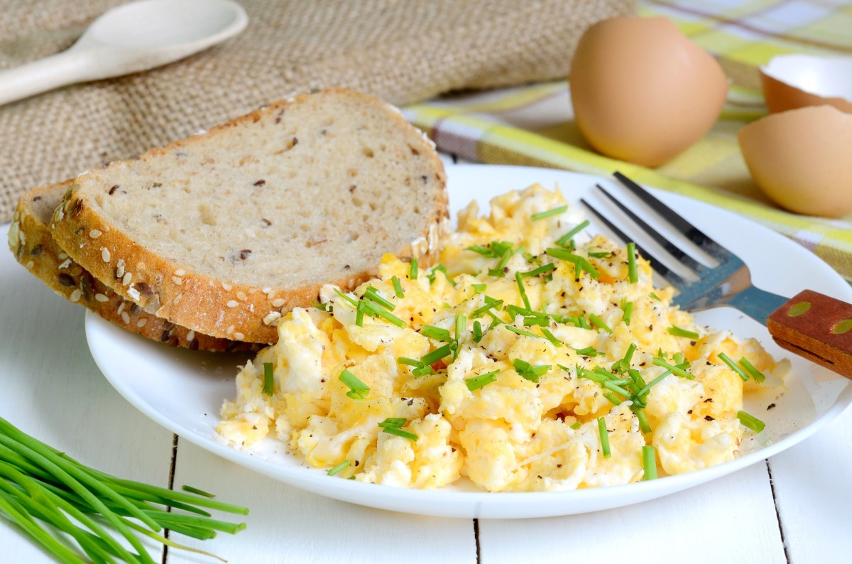 Ein Teller Rührei mit Crème fraîche und zwei Scheiben Brot, daneben Schnittlauch und Eierschalen sowie eine Gabel.