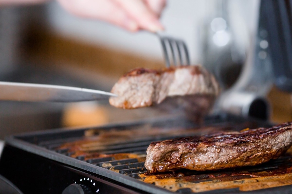 Amazon Black Friday Tefal OptiGrill Kontaktgrill: 2 StÃ¼ck Fleisch auf einem Kontaktgrill, eins wird mit Messer und Gabel weggenommen. Nahaufnahme.