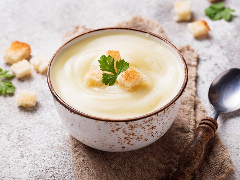 Eine SchÃ¼ssel Apfel-Blumenkohlsuppe mit CroÃ»tons, drumherum weitere CroÃ»tons und ein LÃ¶ffel.