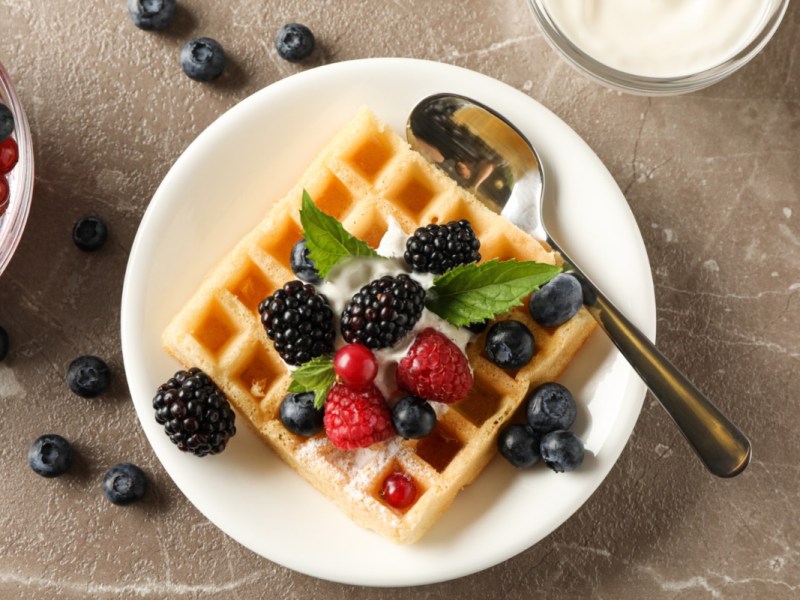 Eine KÃ¤sekuchenwaffel mit FrÃ¼chten, Puderzucker und Sahne auf einem Teller in der Draufsicht.