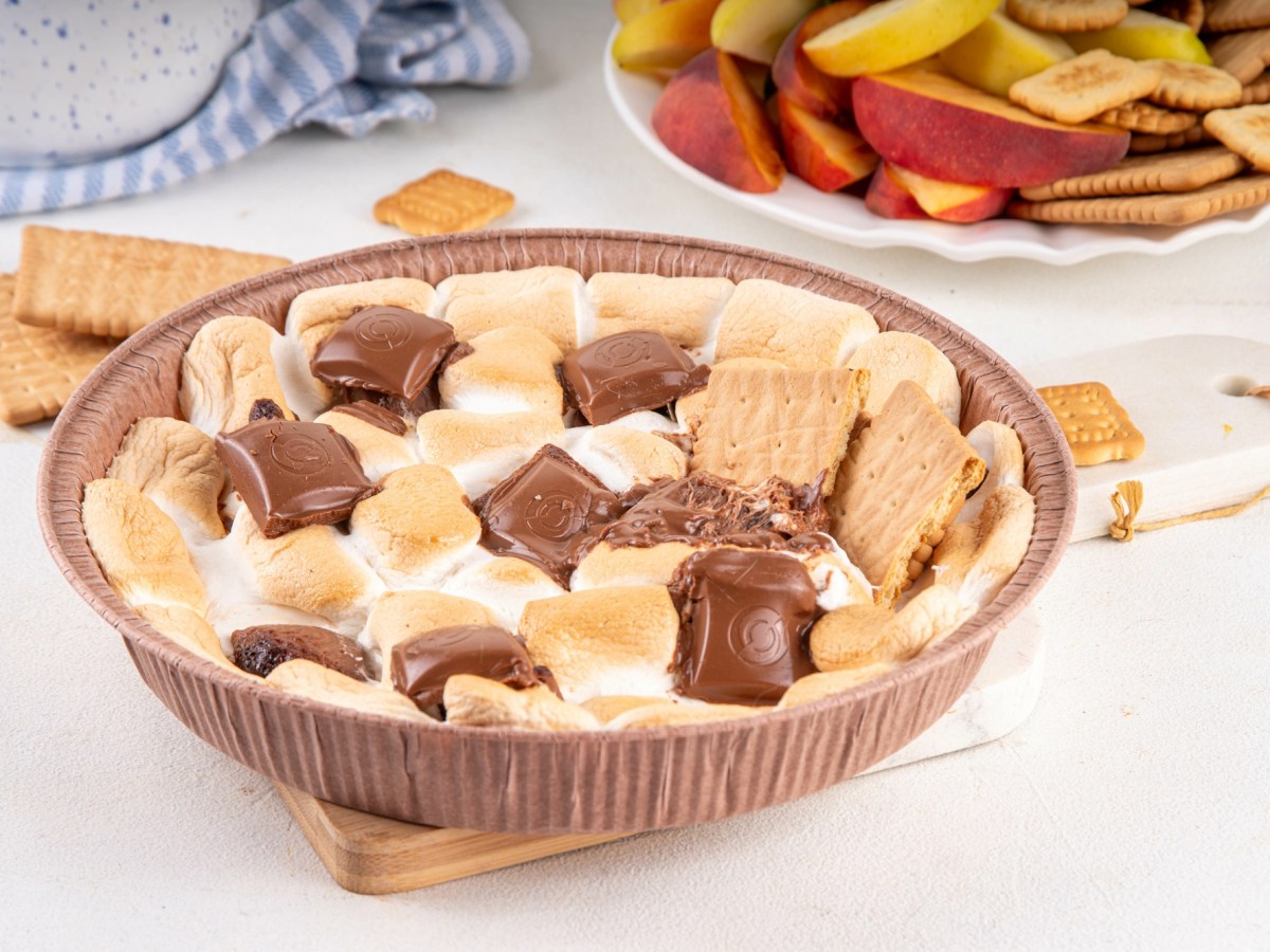 Marshmallow-Creme aus dem Airfryer mit Schokolade und Keksen in einer Pappform, daneben eine Schüssel Obst.