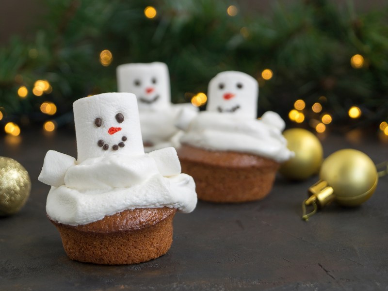 3 Schneemann-Cupcakes mit Marshmallows vor weihnachtlichem Hintergrund.