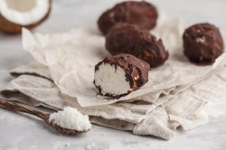 vegane Bounty mit Schokoglasur auf einem Stück Papier, daneben eine halbe Kokosnuss und ein Löffel Kokosraspeln.