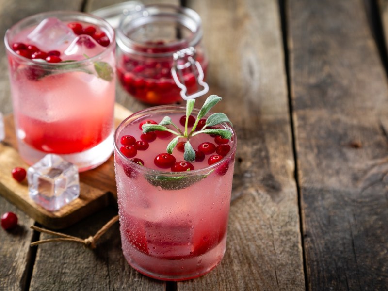 Zwei Gläser Cranberry-Salbei-Mocktail. Im Hintergrund steht ei Einmachglas mit Cranberrys darin.