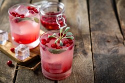 Zwei Gläser Cranberry-Salbei-Mocktail. Im Hintergrund steht ei Einmachglas mit Cranberrys darin.