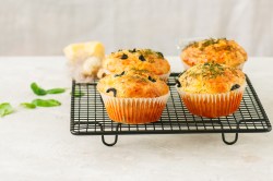 Herzhafte Haferfluckenmuffins mit Kräutern und schwarzen Olivenringen stehen auf einem schwarzen Gitter angerichtet. Im Hintergrund liegen ein paar frische Basilikumblätter.