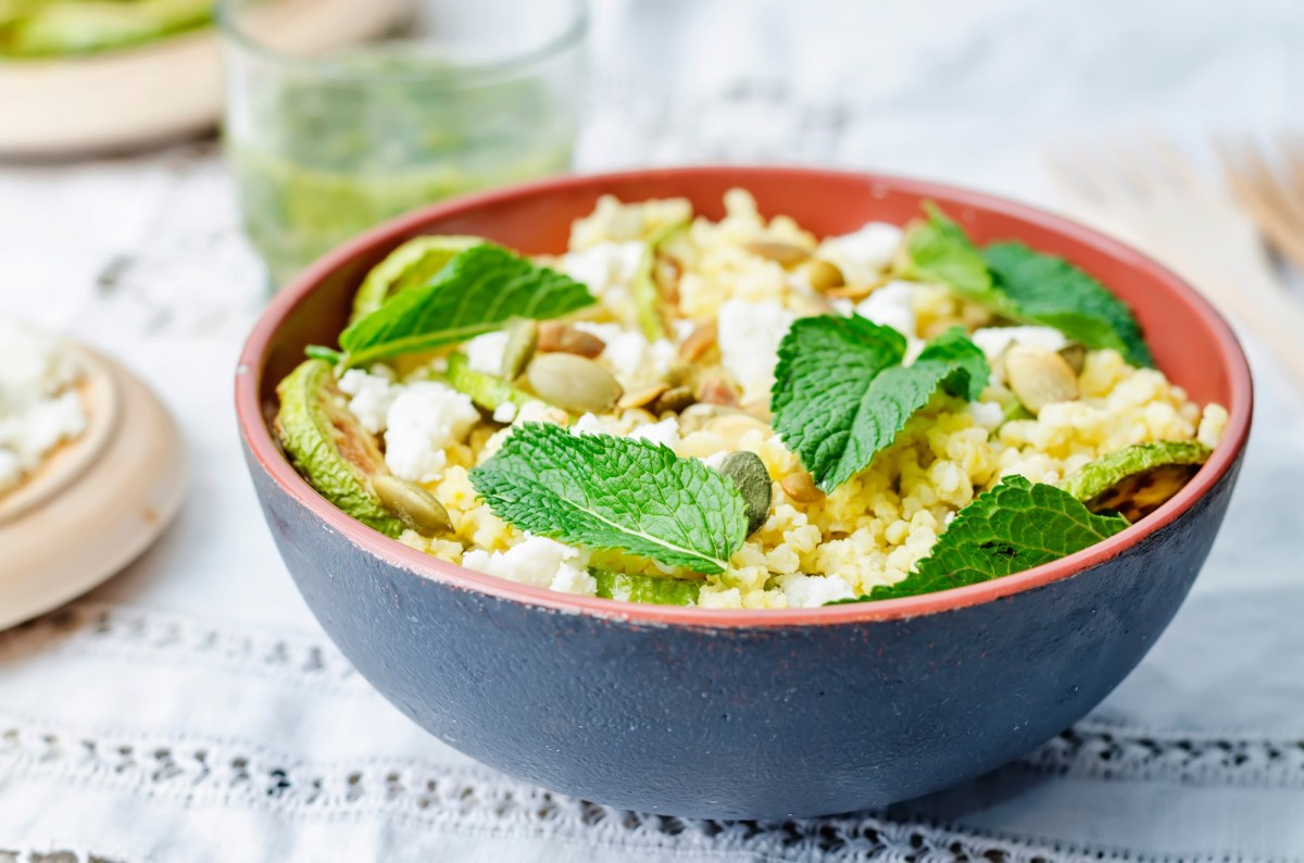 Hirsesalat mit Ziegenkäse in einer . Angerichtet mit frischen Minzblättern in einer schwarz-roten Schüssel auf einem weißen Tuch.