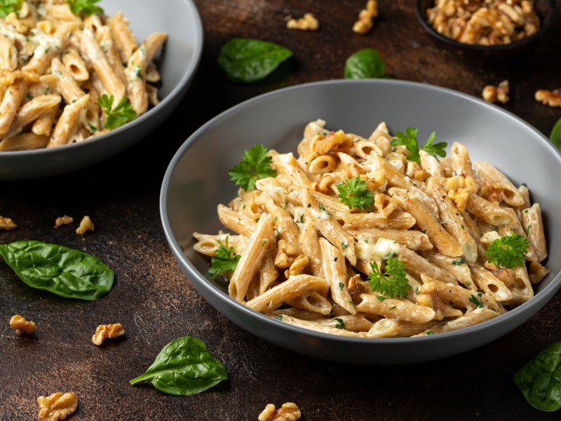 Ein grauer Teller mit einer groÃŸen Portion Hummus-Pasta, die mit frischer Petersilie und gehackten WalnÃ¼ssen serviert wird. Im Hintergrund ist eine weitere Portion des Gerichts zu sehen. Auf dem Tisch liegen BasilikumblÃ¤tter und WalnussstÃ¼ckchen verteilt.