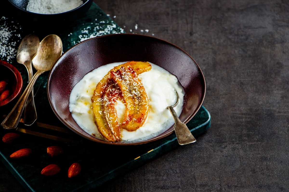 Eine SchÃ¼ssel voll Joghurt mit karamellisierten Bananen. Garniert mit Kokosraspel. Daneben liegen LÃ¶ffel und ein paar Gojibeeren.