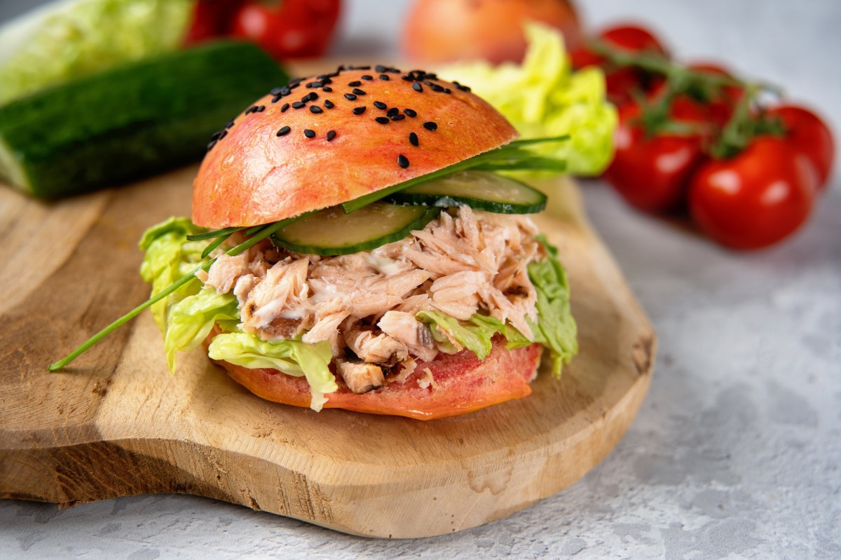 Ein Lachs-Burger mit Gurken und Salat auf einem Brett, im Hintergrund frische Zutaten wie Tomaten.