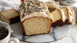 Ein London Fog Cake mit hellem Butterfrischkäse-Frosting und Earl Grey Tee bestreut. Der Kuchen ist kastenförmig und liegt angeschnitten auf einer Marmorplatte. Rechts daneben liegen ein paar weitere Scheiben London Fog Cake. Links auch eine Scheibe. Außerdem ist in der linken unteren Bildecke eine Tasse mit Earl Grey Teemischung zu sehen.