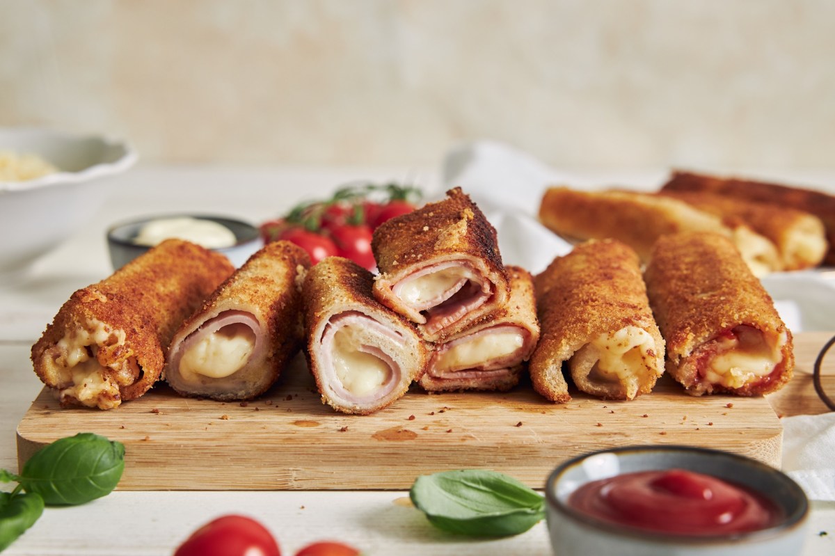 Sieben panierte Toaströllchen, gefüllt mit Schhinken und Käse liegen auf einem Holzbrett. Im Hintergeund liegen weitere Röllchen, Tomaten und Mayonnaise. Im Vordergrund sieht man ein paar Basilikumblätter, eine Schale mit Ketchup und zwei Tomaten.