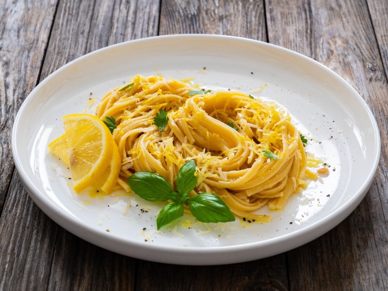 Eine Portion Pasta mit Zitrone und brauner Butter auf einem weiÃŸen, groÃŸen Teller, der auf einem Holztisch steht. Dekoriert ist die Pasta mit Zitrone und brauner Butter mit Zitronenscheiben, frischem Basilikum und geriebenem Parmesan.