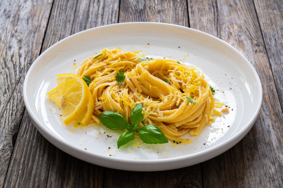 Eine Portion Pasta mit Zitrone und brauner Butter auf einem weiÃŸen, groÃŸen Teller, der auf einem Holztisch steht. Dekoriert ist die Pasta mit Zitrone und brauner Butter mit Zitronenscheiben, frischem Basilikum und geriebenem Parmesan.