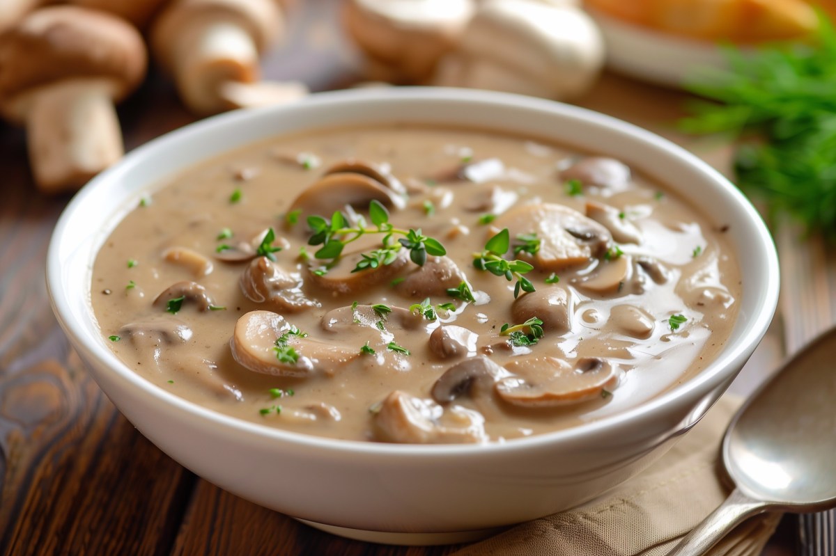 Eine SchÃ¼ssel voll cremigem Pilzeintopf mit weiÃŸen Bohnen, garniert mit frischen KrÃ¤utern. Daneben ein LÃ¶ffel. Im Hintergrund kann man Champignons und anderes GemÃ¼se auf dem Holztisch erkennen.