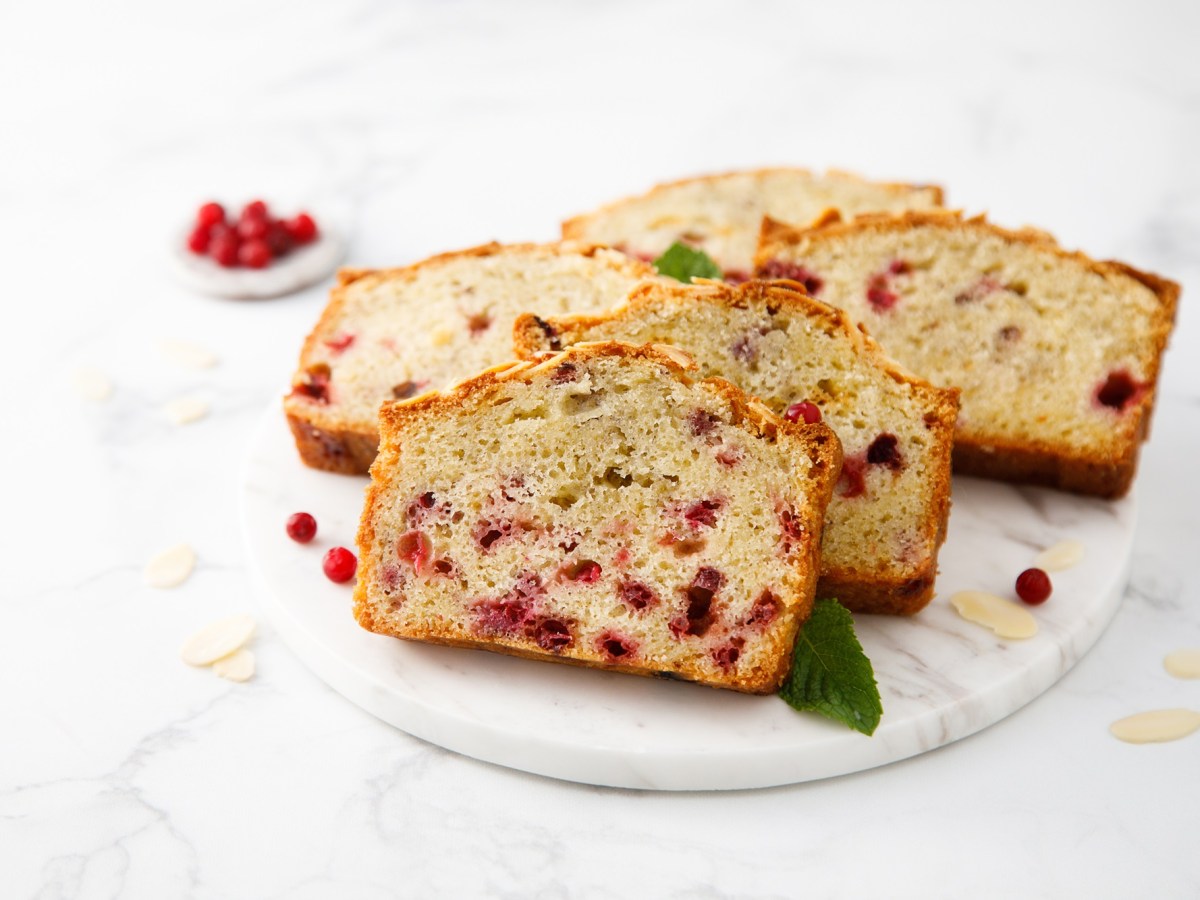 Eine heller marmorner Teller mit fÃ¼nf Scheiben Preiselbeerkuchen. Als Garnitur liegen frische MinzblÃ¤tter dabei. Ringsum liegen einige Mandelsplitter und frische Preisebeeren. Im Hintergrund ist ein kleines SchÃ¤lchen mit Preiselbeeren zu sehen.