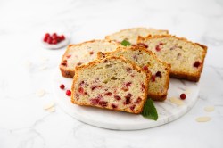 Eine heller marmorner Teller mit fünf Scheiben Preiselbeerkuchen. Als Garnitur liegen frische Minzblätter dabei. Ringsum liegen einige Mandelsplitter und frische Preisebeeren. Im Hintergrund ist ein kleines Schälchen mit Preiselbeeren zu sehen.