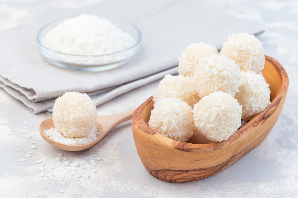 Eine Holzschale mit Raffaello-Energy-Balls auf hellem Untergrund. Im Hintergrund ist eine Glasschale mit Kokosraspel zu sehen, die auf einem hellgrauen Tuch steht. Neben der Holzschale liegt ein HolzlÃ¶ffel mit Kokosraspel und einem einzelnen Raffaello-Energy-Ball.