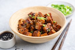 Teriyaki Tofu aus dem Airfryer in Würfel geschnitten in einer kleinen Bambusschale. Sie sind mit Frühlingszwiebeln und hellem sowie schwarzem Sesam angerichtet. Im Hintergrund steht ein kleiner Teller mit geschnittenen Frühlingszwiebeln. Im Vordergrund steht eine Schale mit Sojasoße.