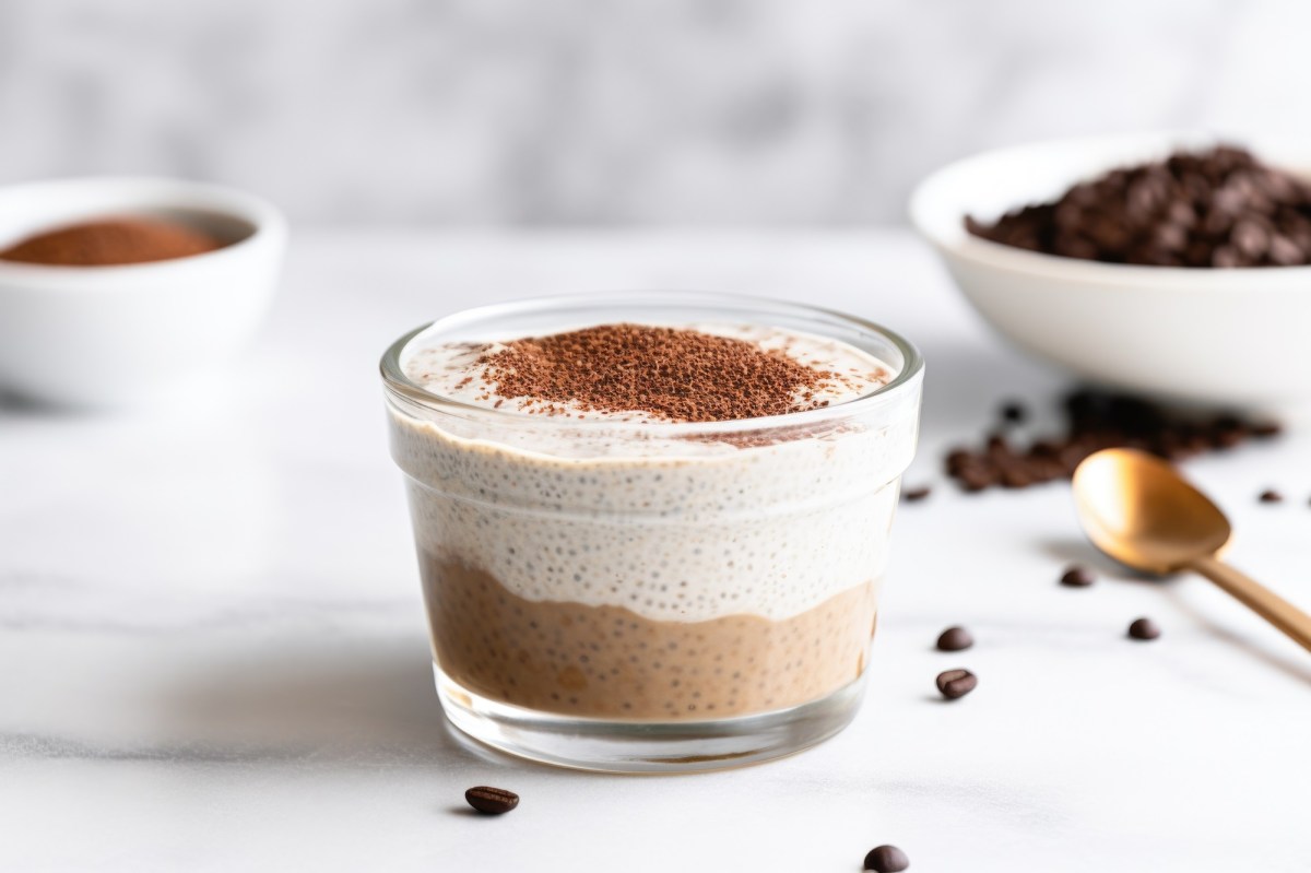 Eine Glasschale mit Tiramisu-Chia-Pudding, bestehend aus einer dunklen Kaffee-Schokoschicht und einer hellen Vanilleschicht steht auf einer marmornen ArbeitsflÃ¤che. Der Tiramisu-Chia-Pudding ist mit Kakaopulver bestreut. Im Hintergrund steht eine weiÃŸe Schale mit Kaffeebohnen und eine kleine Schale mit Kakaopulver. AuÃŸerdem sind ein paar Kaffeebohnen verstreut.