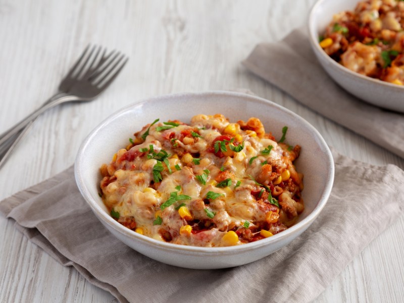 Ein weiÃŸer Teller voll mit veganer One Pot Taco Pasta, der auf einer grauen Stoffserviette steht. SchrÃ¤g links daneben liegen zwei Gabeln. In der rechten oberen Bildecke kann mit verschwommen einen weiteren Teller mit veganer One Pot Taco Pasta erkennen.