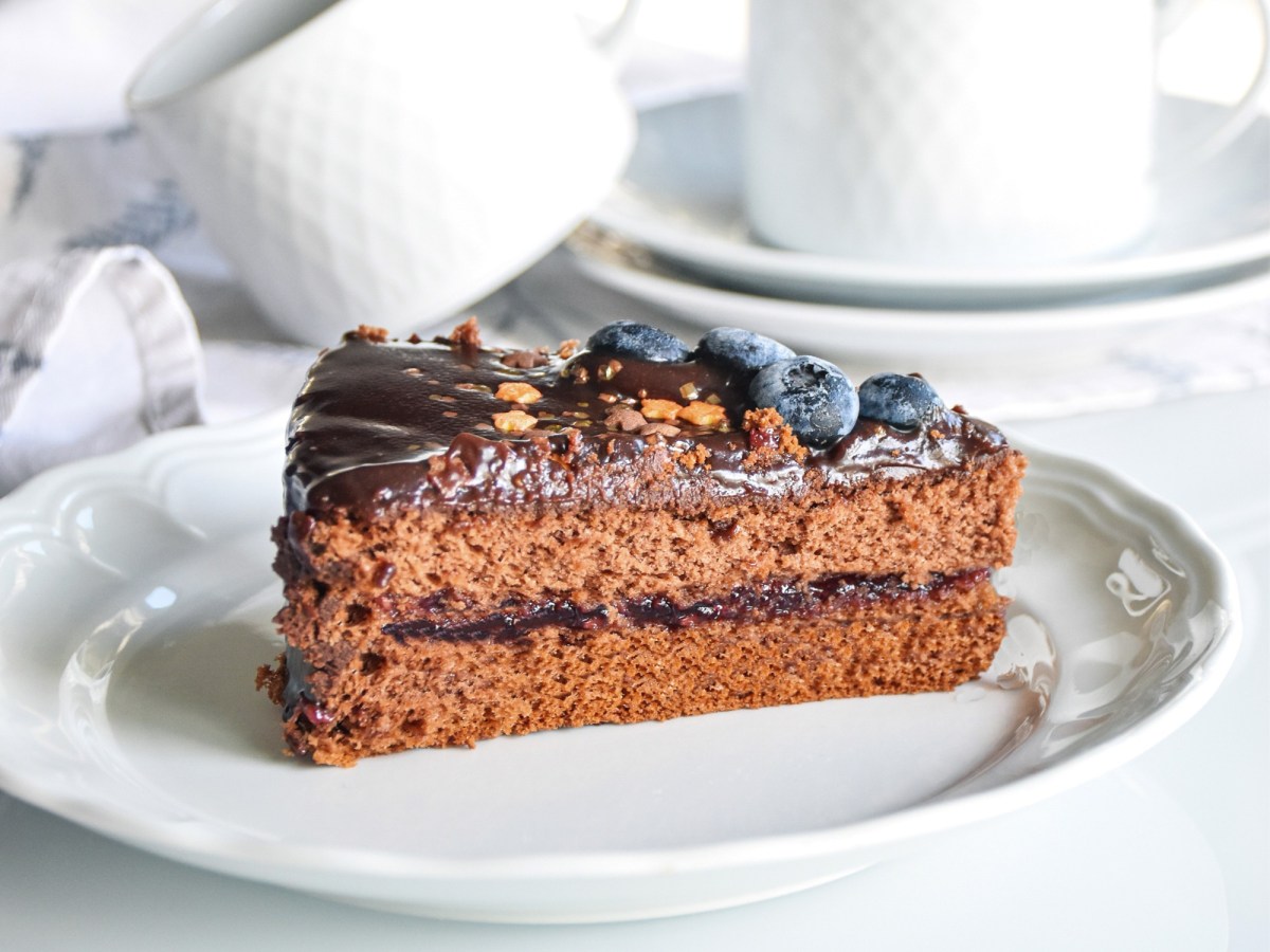 Ein StÃ¼ck vegane Sachertorte auf einem Teller, dahinter Kaffeetassen.