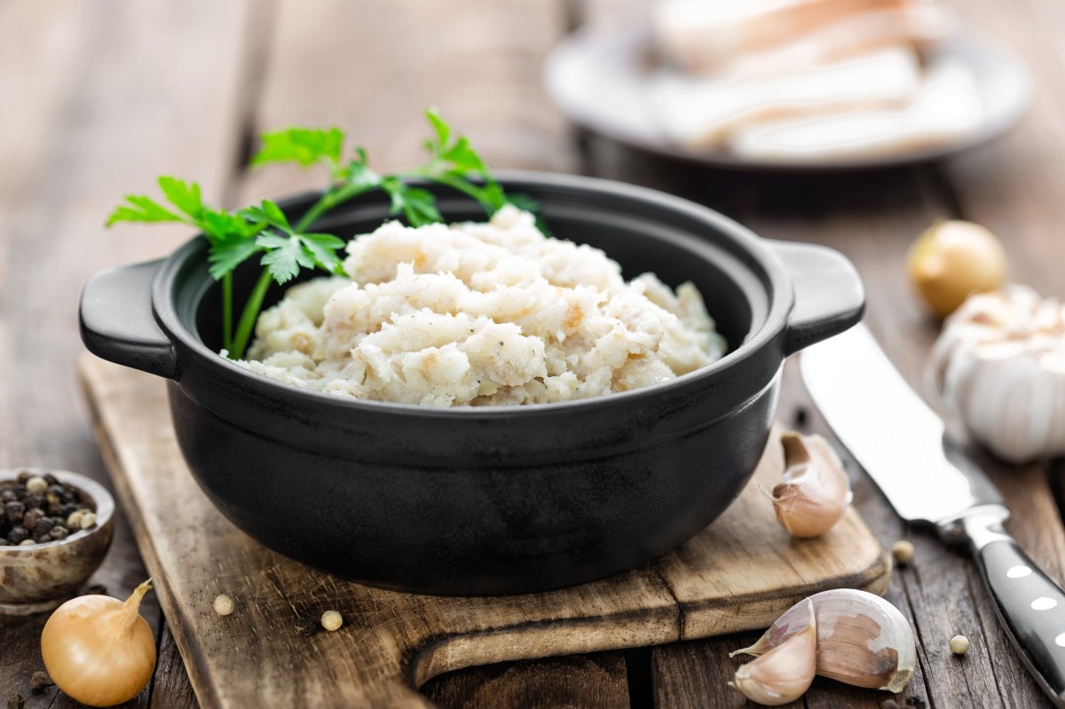 Ein schwarzes TÃ¶pfchen mit veganem Schmalz, garniert mit frischer Petersilie, angerichtet auf einem Holzbrett. Daneben liegt ein Messer, sowie Knoblauchzehen und PfefferkÃ¶rner.