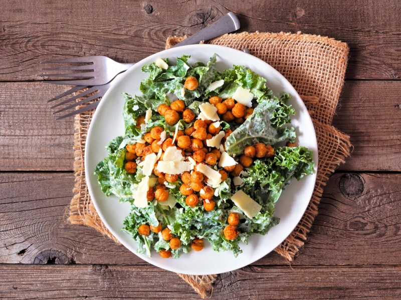 Draufsicht auf einen weiÃŸen Teller mit einer Portion vegetarischem Ceasar Salad mit gerÃ¶steten Kichererbsen und Parmesanflocken. Der Teller steht auf einem Holztisch auf einer hellbraunen rustikalen Stoffserviette. Daneben liegen zwei Gabeln.