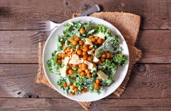 Draufsicht auf einen weißen Teller mit einer Portion vegetarischem Ceasar Salad mit gerösteten Kichererbsen und Parmesanflocken. Der Teller steht auf einem Holztisch auf einer hellbraunen rustikalen Stoffserviette. Daneben liegen zwei Gabeln.