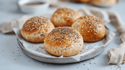 Ein Teller mit weißem Backpapier auf dem vier Dinkel-Skyr-Brötchen liegen, die mit Sesam und Chiasamen bestreut sind. Im Hintergrund kann man verschwommen eine Schale und weitere Brötchen erkennen. Um den Teller liegt ein beiges Küchentuch.