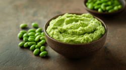 Eine Schale gefüllt mit Edamame-Hummus. Links neben der Schale liegen Edamamebohnen. Im Hintergrund steht eine weitere Schale mit Edamamebohnen.