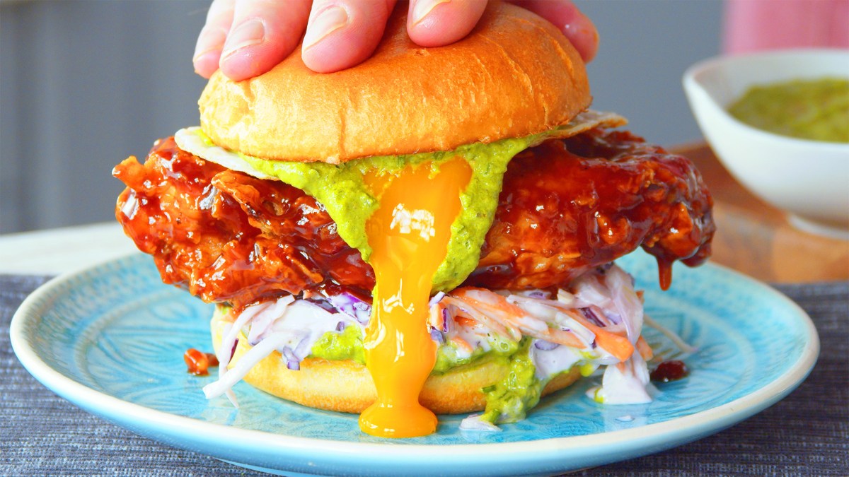 Ein Fried Chicken Burger auf einem blauen Teller wird leicht zerdrückt, sodass Soßen und Eigelb etwas aus ihm herauslaufen.